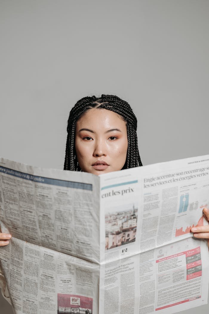 Asian woman with braided hair reading a newspaper against a white background.